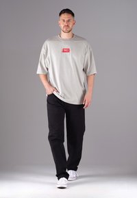 Helles graues T-Shirt mit kurzen Ärmeln und rotem Logopatch, kombiniert mit schwarzen Hosen und weißen Sneakers. Das Shirt hat einen lässigen Schnitt und seitliche Schlitze.