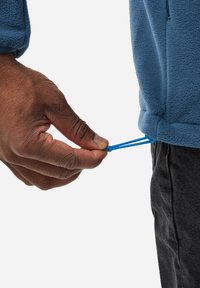 Blue fleece jacket with a drawstring hem. A hand pulls the blue drawstring while wearing dark jeans. The texture of the fabric is visible.