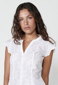 Blouse blanche brodée de fleurs avec des manches courtes à bords festonnés, un devant à boutons et des détails en broderie ajourée sur l'ensemble du tissu.