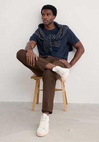 Jūrinės spalvos marškiniai, rudi džinsai, kreminės spalvos aukšti sportbačiai. Vilnos ir poliesterio šalikėlis su mėlynomis ir baltomis juostelėmis užmestą ant pečių. Sėdi ant medinės taburetės.