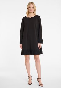 Schwarzes Kleid mit Rundhalsausschnitt, langen Ärmeln und fransigen Säumen, die mit Perlen verziert sind. Glatter Stoff und lässige Passform.