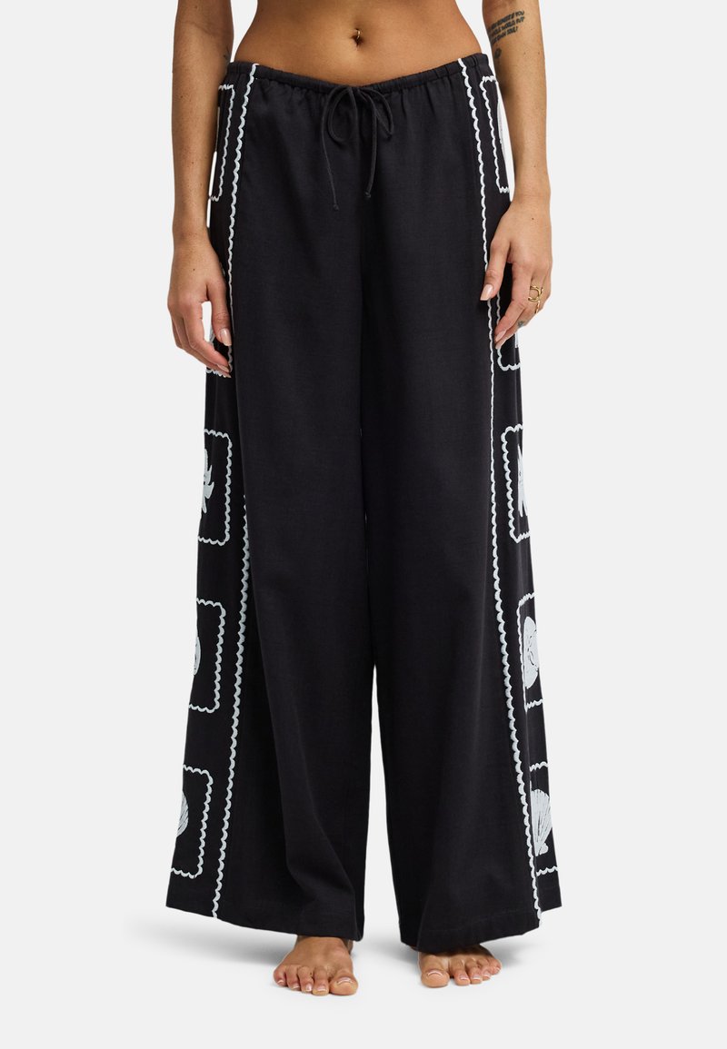 Μαύρο παντελόνι με φαρδιά παντελόνια με λευκά μοτίβα κοχυλιών και κυματιστά τετράγωνα σχέδια στα πλαϊνά, φορεμένο από ξυπόλητο άτομο με ορατό τατουάζ στο χέρι.