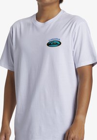 Camiseta de algodón blanca con mangas cortas. Presenta un logotipo colorido de Quiksilver en el lado izquierdo del pecho con un diseño circular. Textura suave.