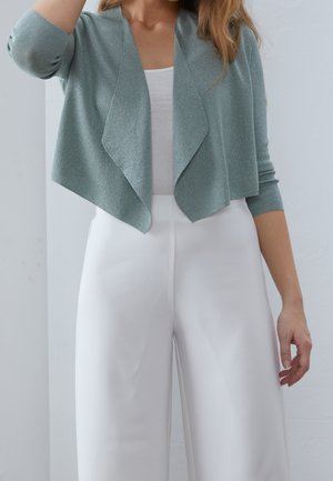 Femme portant un cardigan bleu clair ouvert sur un top blanc, associé à un pantalon blanc taille haute à jambes larges.