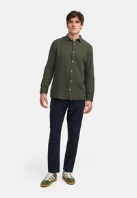 Olivgrünes, langärmeliges Hemd mit strukturiertem Muster, Knopfleiste, kombiniert mit dunkelblauen Hosen und grünen Sneakers mit weißen Akzenten.