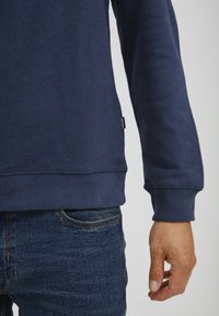Navy blue sweatshirt with ribbed hem and cuffs, featuring a subtle logo tag on the side, paired with denim jeans.