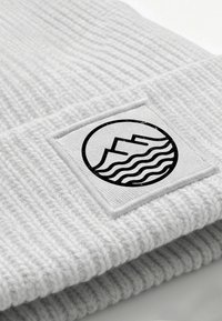 White ribbed knit beanie with a square patch. The patch features a black circular design with waves and mountains. Soft texture, casual design.