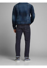 Denimjacka i mörkblått, med urtvättad textur och sömdetaljer, kombinerad med mörka byxor och grå mockasneakers, sedd bakifrån.