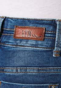 Traperice s smešnim kožnim štepom na kojem je prikazano "LTB" i logo valova; plava tkanina s vidljivim šavovima i blagom iznošenosti.