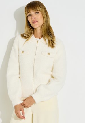 Mujer con cabello largo y rubio que lleva una chaqueta blanca y esponjosa con cremallera y botones dorados en los bolsillos del pecho, sobre un fondo claro y liso.