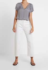 Top de satén gris con escote en V y detalles de encaje, combinado con jeans rectos blancos y sandalias negras de tiras, de pie frente a un fondo neutro.
