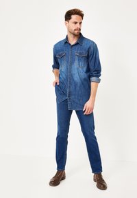 Denimhemd mit zwei Brusttaschen, hochgekrempelten Ärmeln und lockerer Passform, kombiniert mit blauen Jeans und braunen Schnürstiefeln.