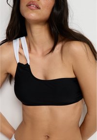 Fekete, egyvállas bikini felső fehér pánttal. Simító, rugalmas anyag jellemzi, és a vállnál gyűrődik, hogy biztos illeszkedést nyújtson.