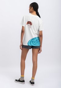 T-shirt bianca in cotone con grafica colorata di un paracadute sulla schiena e un design circolare blu vicino all'orlo. Maniche corte, vestibilità morbida, indossato con pantaloni corti grigi.