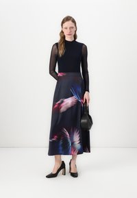 Haut noir à manches longues, manches transparentes, jupe maxi à motif floral dans des tons foncés. Chaussures noires unies et sac à main structuré.