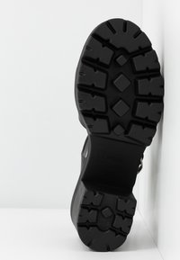 Semelle de chaussure en caoutchouc noir présentant un motif de semelle robuste avec des rainures profondes et des sections inclinées pour l'adhérence. Surface lisse autour du talon.
