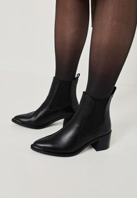 Černé kožené kotníkové boty s špičatou špičkou, elastickými bočními panely a masivním podpatkem. Hladký povrch a jednoduché designové detaily.