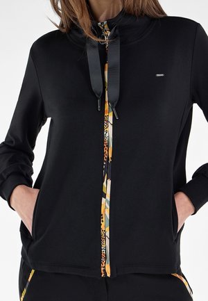 Person mit einer schwarzen Jacke mit Reißverschluss, der bunte Muster an Reißverschluss und Taschen aufweist, Hände in den Taschen verstaut.