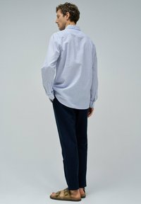 Camisa de rayas azul y blanca de mangas largas, de corte holgado, combinada con pantalones oscuros y sandalias marrones. Tejido suave, costuras y botones visibles.