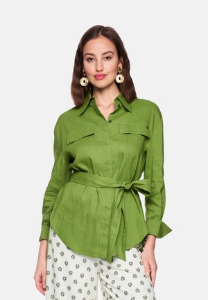 Woman wearing a green belted button-up shirt with long sleeves and white pants with a small floral pattern, standing against a plain background.