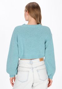 Frau mit hellbraunen Haaren, die einen kurz geschnittenen hellblauen Strickpullover und hochgeschnittene helle Denim-Jeans trägt, mit dem Rücken zur Kamera.