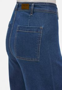 Jeans de mezclilla azul oscuro con un bolsillo trasero, costuras naranjas y una etiqueta marrón en la cintura que presenta la marca "YSABEL MORA".