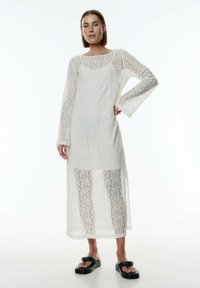 Langes, weißes Spitzenkleid mit langen Ärmeln und einem durchsichtigen Überwurf. Verfügt über einen runden Ausschnitt und ein aufwendiges florales Spitzenmuster. Kombiniert mit schwarzen Sandalen.