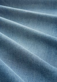 Svetlo modra-denim tkanina z gladko teksturo, ki prikazuje gube in subtilen lesk ter klasične enosmerne vzorce. 