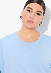 Maglietta azzurra chiara a maniche corte con scollatura rotonda e texture morbida. Il modello indossa orecchini a cerchio in argento, con uno sfondo minimale.