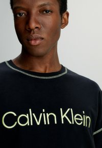 Černé tričko s limetkově zelenou výšivkou s nápisem "Calvin Klein", se kontrastním prošíváním kolem krku a hladkým bavlněným povrchem.