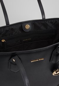 Sac fourre-tout en cuir noir avec intérieur matelassé, garnitures dorées et poche zippée. Comprend un logo embossé et deux sangles d'épaule.