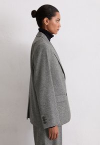 Blazer en laine grise avec une coupe cintrée, un col cranté, des poches latérales et des poignets à trois boutons, porté avec un col roulé noir en dessous.