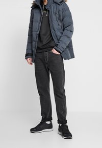 Chaqueta acolchada de color azul oscuro con capucha con borde de piel, combinada con una sudadera negra y vaqueros gris oscuro, completada con zapatillas negras.