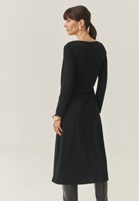 Rochie neagră cu mâneci lungi, decolteu rotund, croi slim și lungime până la genunchi. Textură netedă, design minimal, completată cu o brățară aurie.