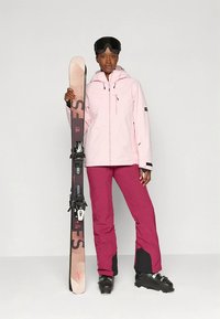 Rózsaszín vízálló sídzseki kapucnival, bordó sínadrággal párosítva. Síléceket tart, melyeken kötés van, fekete sícipőben. Hóvégzésű símaszk a fejen.