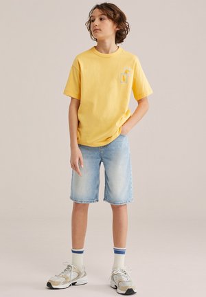 Fant z rjavimi lasmi, oblečen v rumeno majico z grafiko ananasa, lahke jeans hlače, bele nogavice z modrimi črtami in superge.