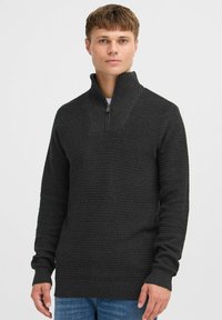 Dunkelgrauer Strickpullover mit hohem Kragen und halbem Reißverschluss, ausgestattet mit gerippter Textur und langen Ärmeln. Getragen über einem weißen Hemd und blauen Jeans.