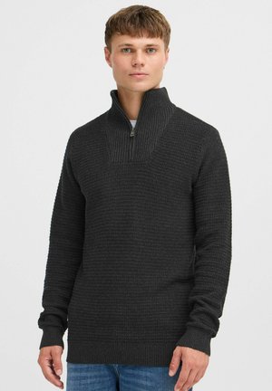 Mørk grå strikkegenser med høy krage og halv glidelås, med ribbestruktur og lange ermer. Brukt over en hvit skjorte og blå jeans.