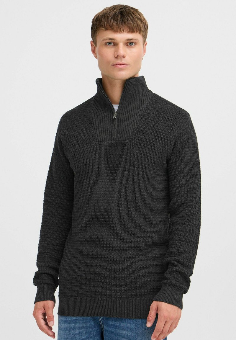 Dunkelgrauer Strickpullover mit hohem Kragen und halbem Reißverschluss, ausgestattet mit gerippter Textur und langen Ärmeln. Getragen über einem weißen Hemd und blauen Jeans.