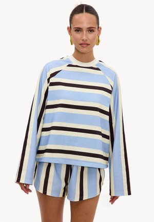 Femme portant un haut ample à manches longues rayé bleu, crème et noir avec un short rayé assorti et des boucles d'oreilles créoles dorées.
