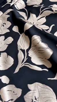 Crni materijal s uzorkom u boji slonovače s detaljnim ljiljanima i lišćem, prikazujući teksturu tkanine i blage nabore.