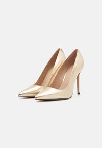 Gullfargede patentlæder høyhælte pumps med spiss tå, en jevn overflate og en tynn stilettohæl, med et lavt vampdesign.