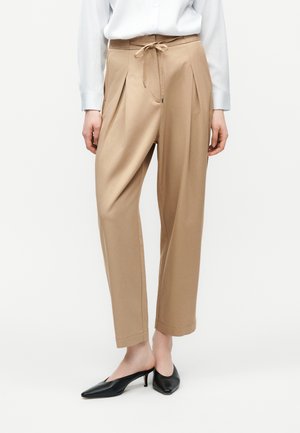 Kvinde iført beige højtsiddende, tilspidsede bukser med pressede folder foran og snøre, kombineret med sorte slingback-sko med lav hæl og spids tå.