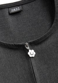 Encolure de vêtement gris foncé avec tirette de fermeture éclair argentée en forme de fleur et étiquette noire portant l'inscription "NEXT Made in Sri Lanka."