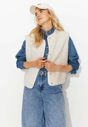 Gilet en polaire crème avec fermeture à pression, superposé sur une chemise en denim. Coupe ample, associé à un jean en denim décontracté, créant un look texturé.