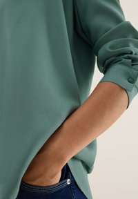 Blouse en soie verte à manches longues, avec une coupe décontractée et des poignets boutonnés. La texture est lisse et elle est partiellement rentrée dans un jean bleu.