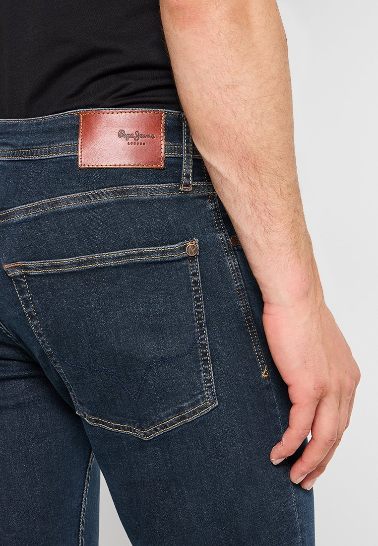 Dunkelblaue Jeans aus Denim mit schlanker Passform, ausgestattet mit einem braunen Lederaufnäher auf der Rückseite und kontrastierenden Nähten. Die Gesäßtasche enthält eine dekorative Naht.