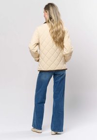 Beežist tikitud jakk teemantmustriga, pruuni äärisega allääres, kantud siniste lõhestatud teksade ja beežide jalanõudega, vaadatuna tagant.