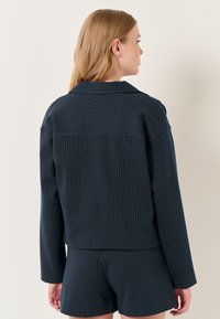 Donkerblauw jasje met krijtstreepjes, een cropped ontwerp, lange mouwen en een klassieke kraag. Zachte stof met fijne witte en gouden strepen.