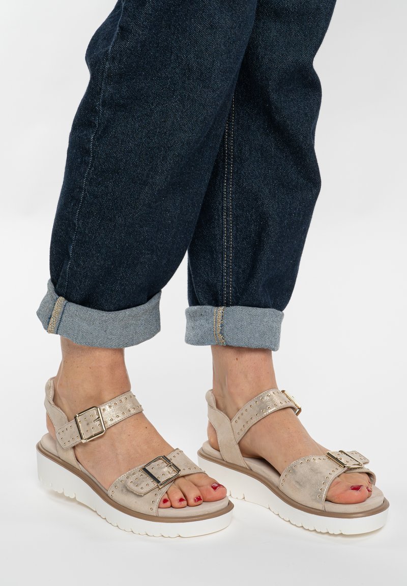 Pieds portant des sandales beiges avec boucles et semelles blanches, associées à un jean denim bleu foncé retroussé, sur un fond uni.